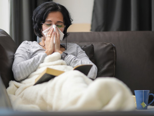 Eine erkältete Frau sitzt unter einer warmen Decke auf der Couch und schnaubt sich die Nase